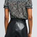 A model shown from the back wearing a short-sleeve black and gray snakeskin print blouse with puff sleeves, paired with black faux-leather shorts, against a light gray studio background.