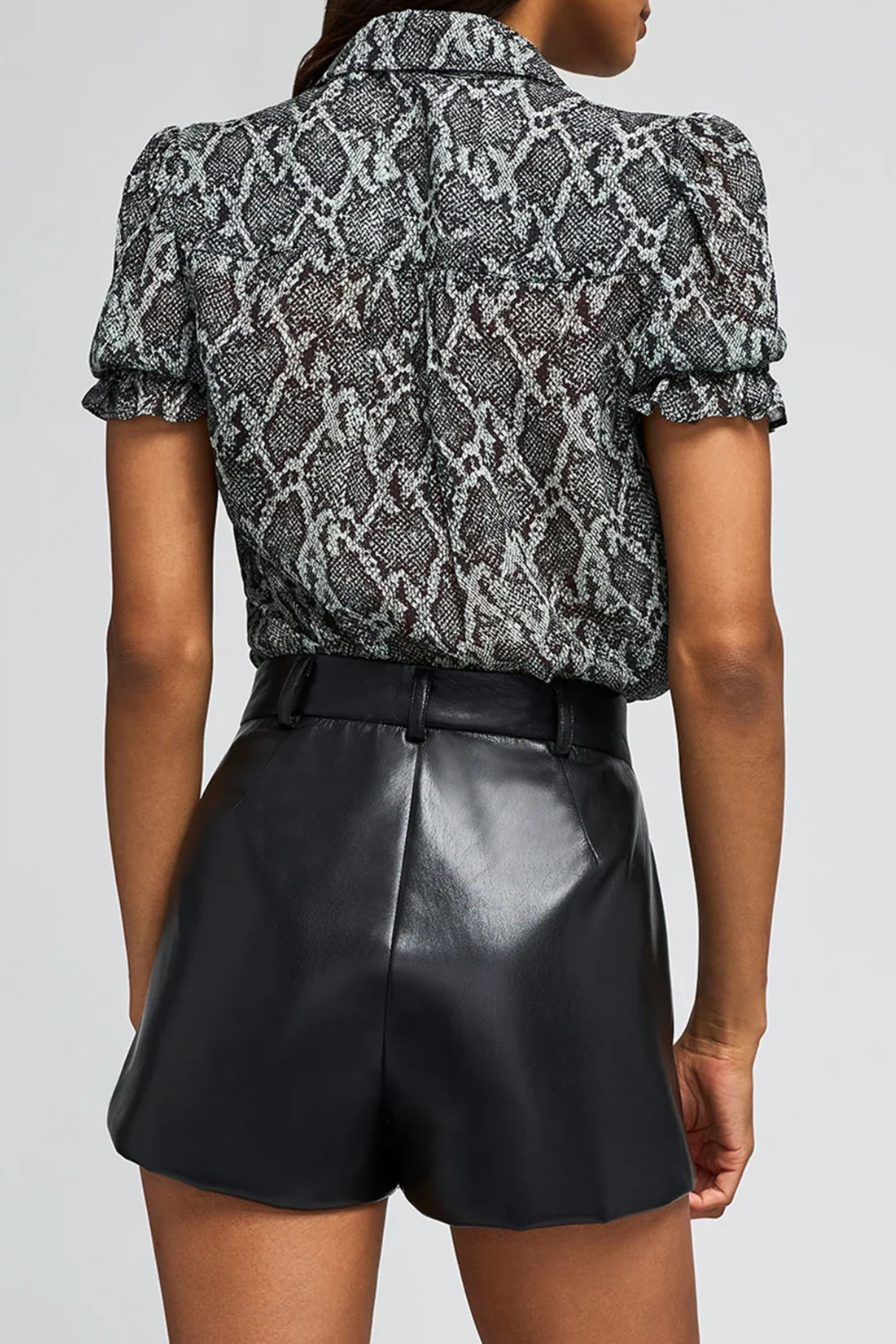 A model shown from the back wearing a short-sleeve black and gray snakeskin print blouse with puff sleeves, paired with black faux-leather shorts, against a light gray studio background.
