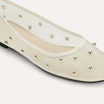 A close-up of a cream-colored ballet flat shoe with metallic gold studs and circular cutouts along the side, set against a solid light beige background.