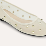 A close-up of a cream-colored ballet flat shoe with metallic gold studs and circular cutouts along the side, set against a solid light beige background.