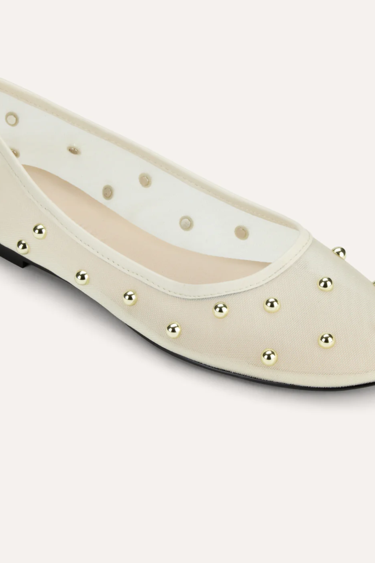 A close-up of a cream-colored ballet flat shoe with metallic gold studs and circular cutouts along the side, set against a solid light beige background.