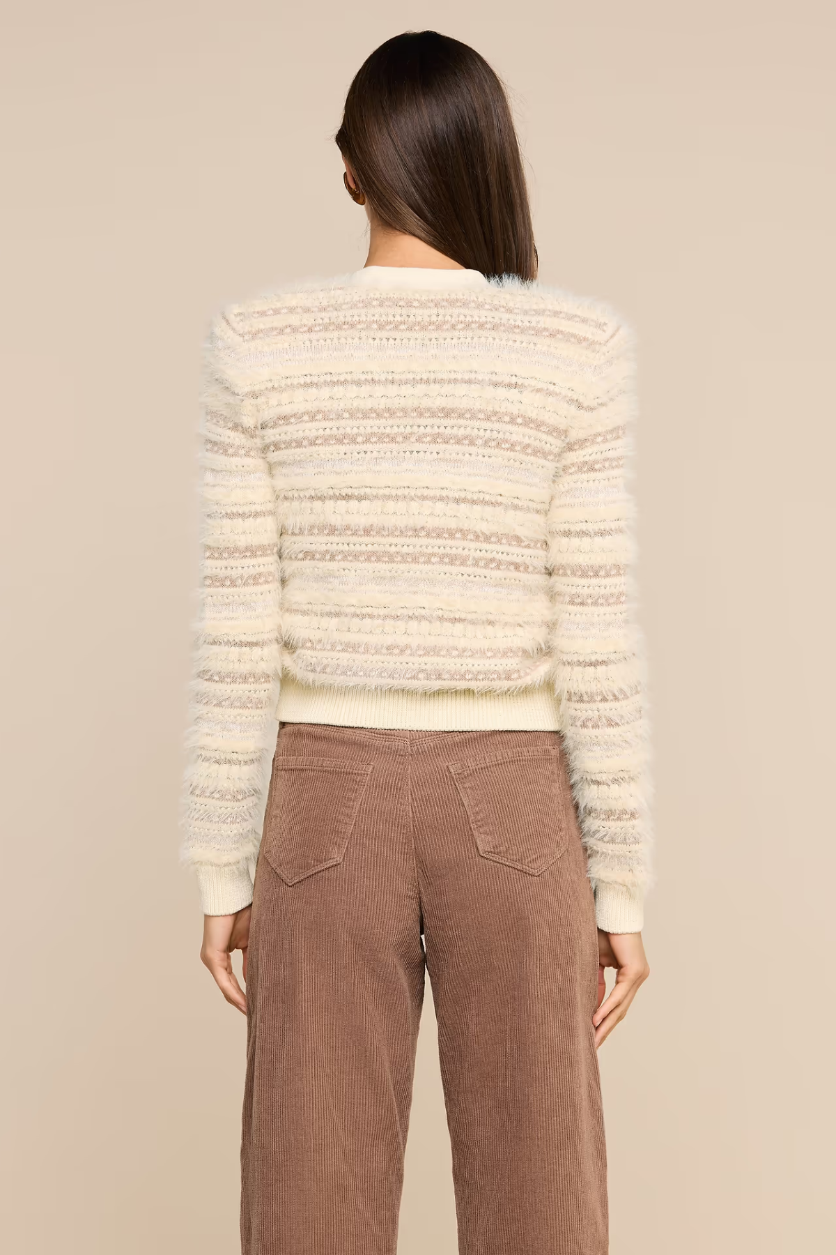 A woman shown from the back wearing a cream-colored textured knit sweater with horizontal striped patterns and brown wide-leg pants, set against a solid beige studio background.