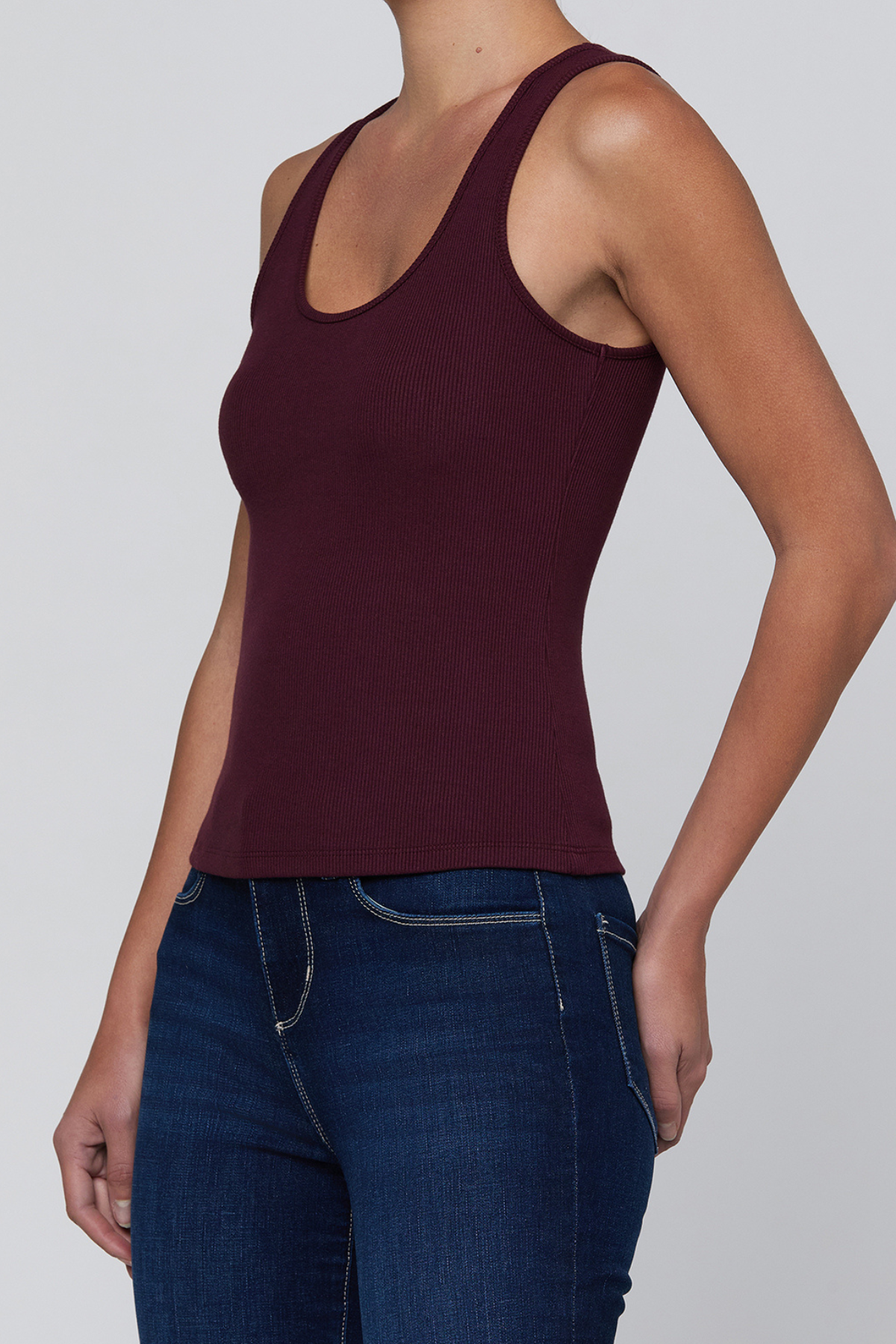 A woman wearing a sleeveless scoop-neck tank top in deep burgundy, paired with dark blue jeans, shown from the neck down against a light gray studio background.