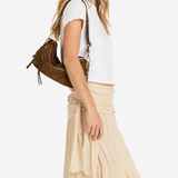 A woman wearing a white short-sleeve crop top and a beige ruffled tiered maxi skirt, carrying a brown leather shoulder bag against a plain white background.