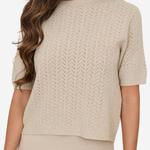 A woman wearing a short-sleeve knit sweater in a light beige or cream color, paired with a matching knit skirt. The sweater features a textured chevron or herringbone knit pattern and a crew neckline.
