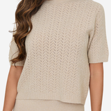 A woman wearing a short-sleeve knit sweater in a light beige or cream color, paired with a matching knit skirt. The sweater features a textured chevron or herringbone knit pattern and a crew neckline.