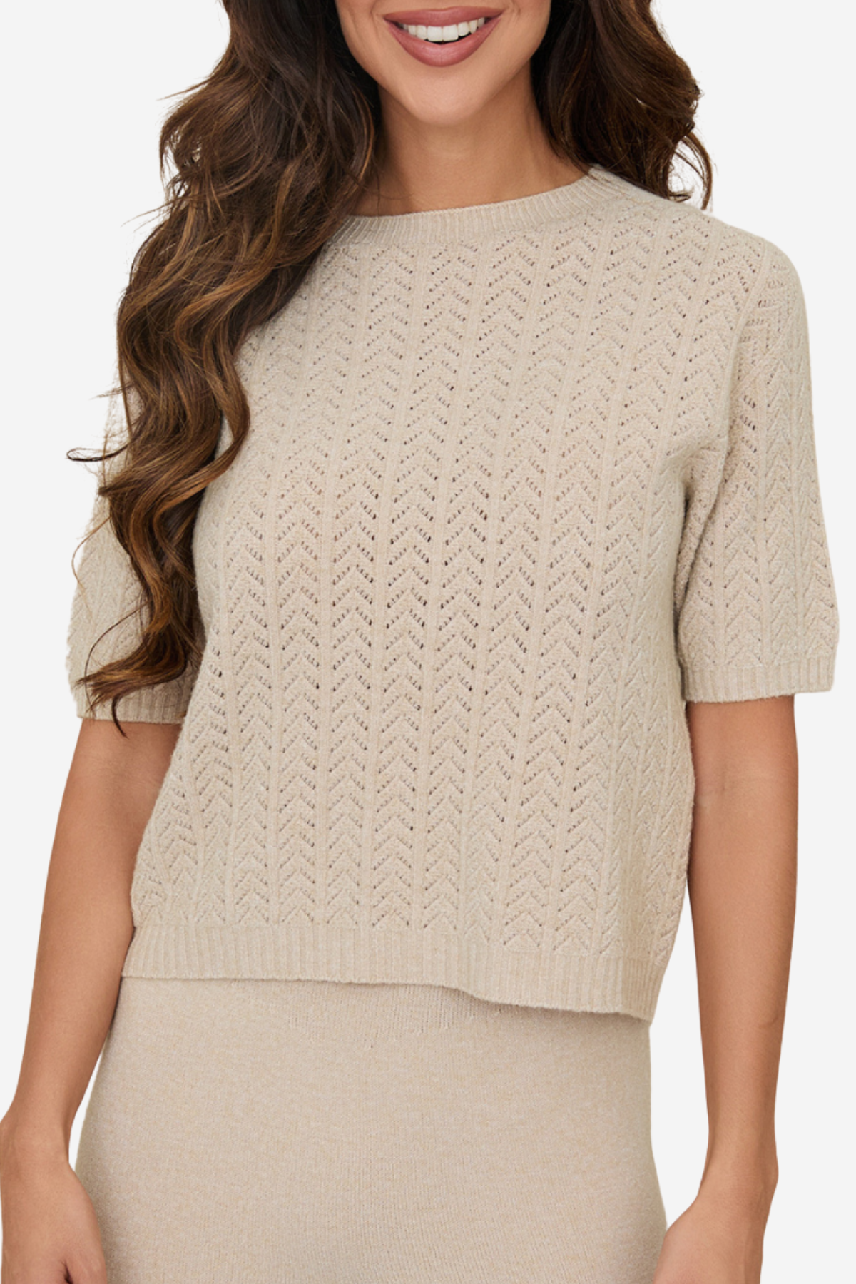 A woman wearing a short-sleeve knit sweater in a light beige or cream color, paired with a matching knit skirt. The sweater features a textured chevron or herringbone knit pattern and a crew neckline.