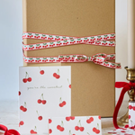 A kraft paper gift box tied with a red and white cherry-patterned ribbon, displayed with matching cherry-print cards and a small gift box on a light marble surface beside a lit candle.