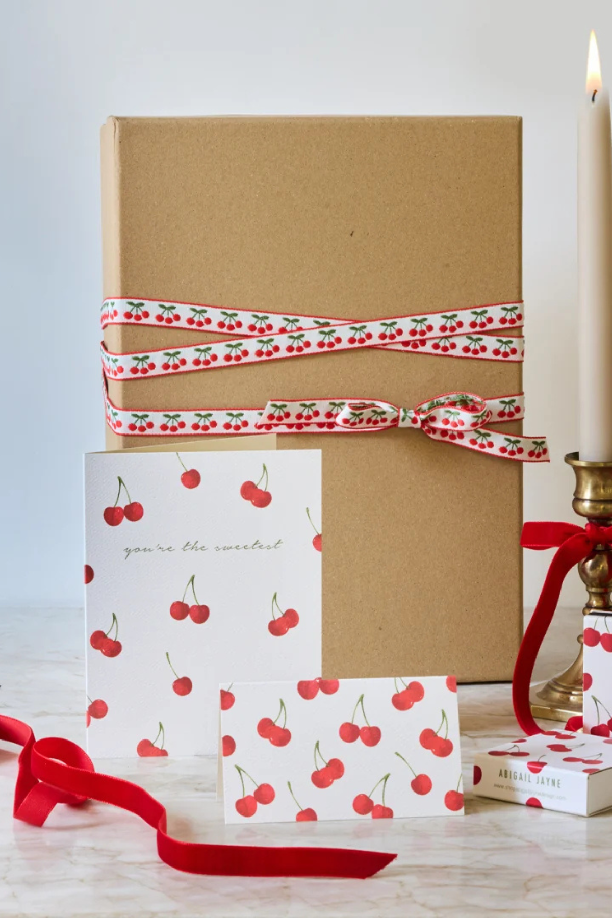 A kraft paper gift box tied with a red and white cherry-patterned ribbon, displayed with matching cherry-print cards and a small gift box on a light marble surface beside a lit candle.