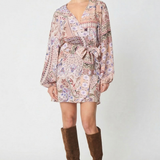 A model wearing a long-sleeved mini wrap dress with a pink and purple paisley pattern, styled with brown suede knee-high boots against a light gray studio background.