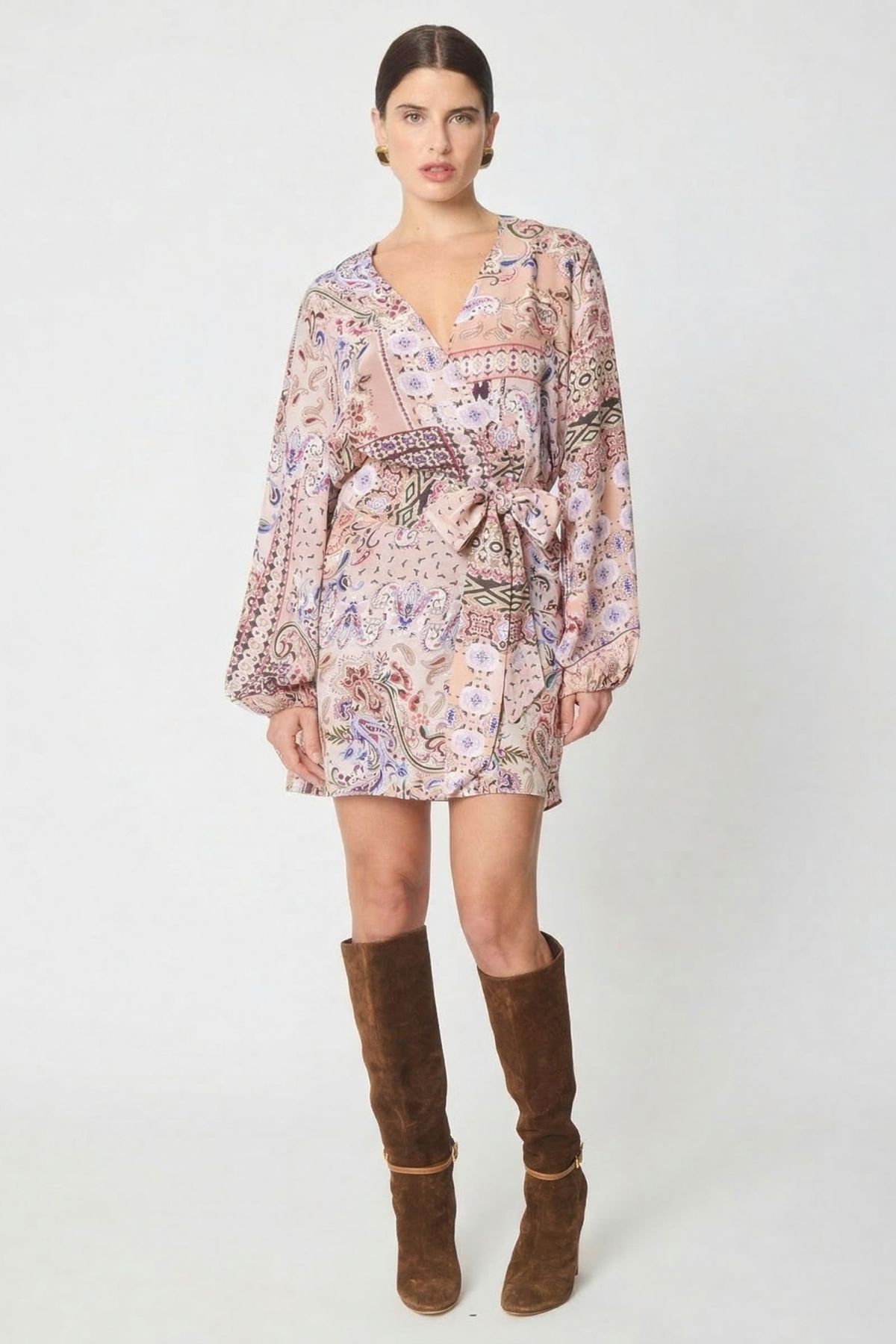 A model wearing a long-sleeved mini wrap dress with a pink and purple paisley pattern, styled with brown suede knee-high boots against a light gray studio background.