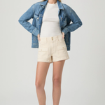 Woman wearing a denim jacket, white shirt, and beige shorts on a plain background