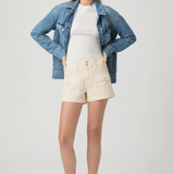 Woman wearing a denim jacket, white shirt, and beige shorts on a plain background