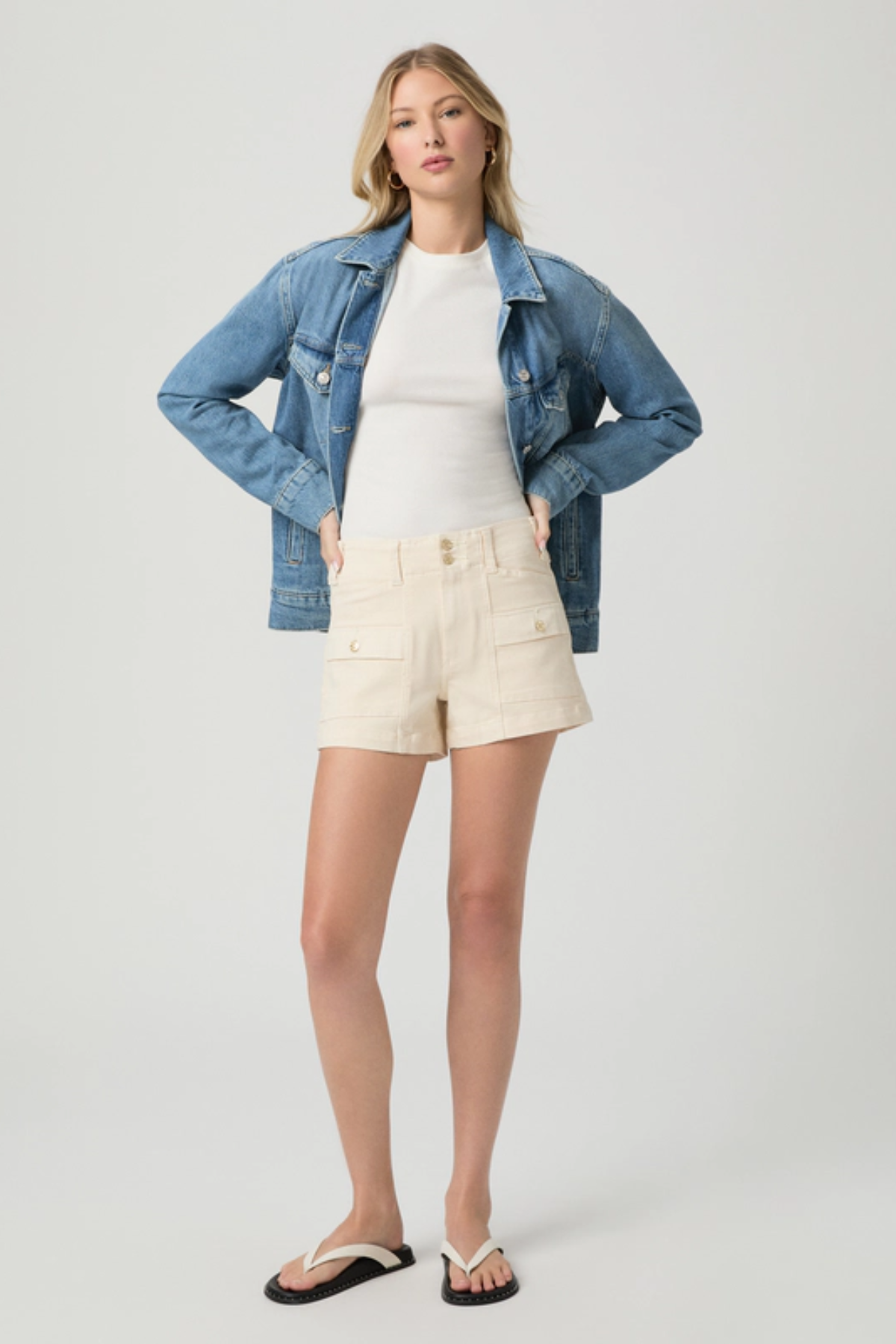 Woman wearing a denim jacket, white shirt, and beige shorts on a plain background