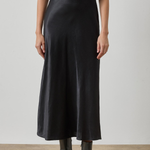 A model wearing a black satin midi skirt with a matching black short-sleeve top and black leather ankle boots, standing against a neutral studio background.