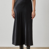 A model wearing a black satin midi skirt with a matching black short-sleeve top and black leather ankle boots, standing against a neutral studio background.