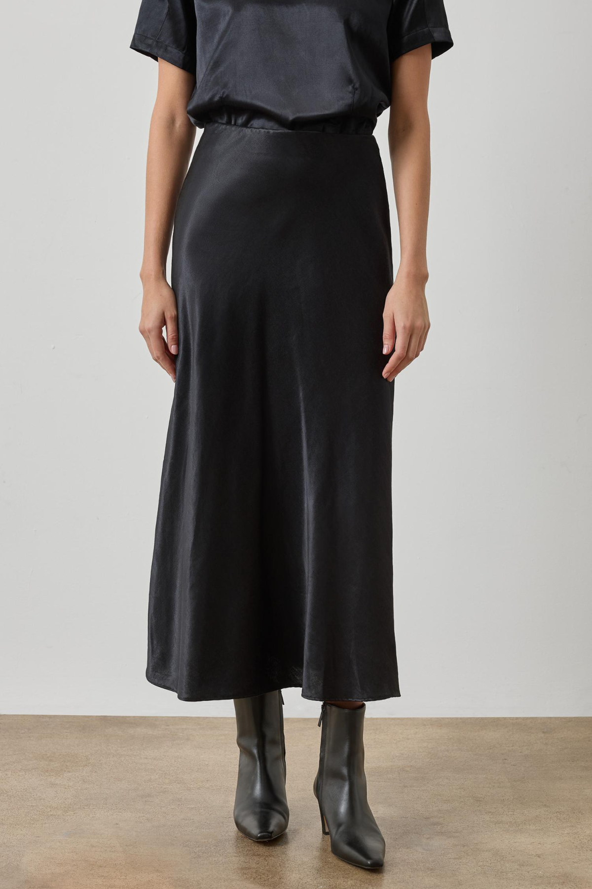 A model wearing a black satin midi skirt with a matching black short-sleeve top and black leather ankle boots, standing against a neutral studio background.