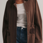 A model wearing a brown corduroy utility jacket over a white tank top and blue jeans, shown from the neck down against a light gray studio background.