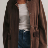 A model wearing a brown corduroy utility jacket over a white tank top and blue jeans, shown from the neck down against a light gray studio background.