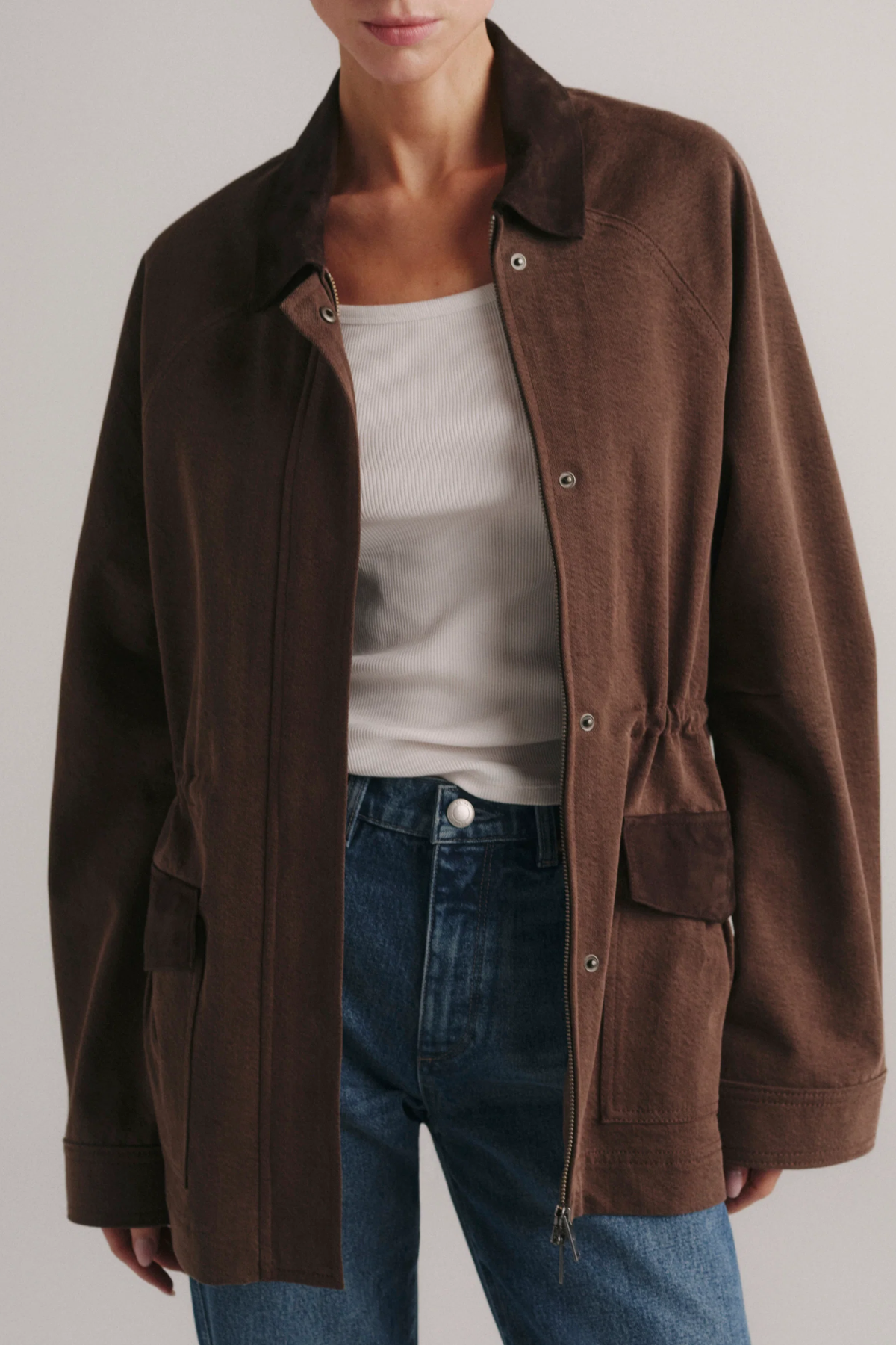 A model wearing a brown corduroy utility jacket over a white tank top and blue jeans, shown from the neck down against a light gray studio background.