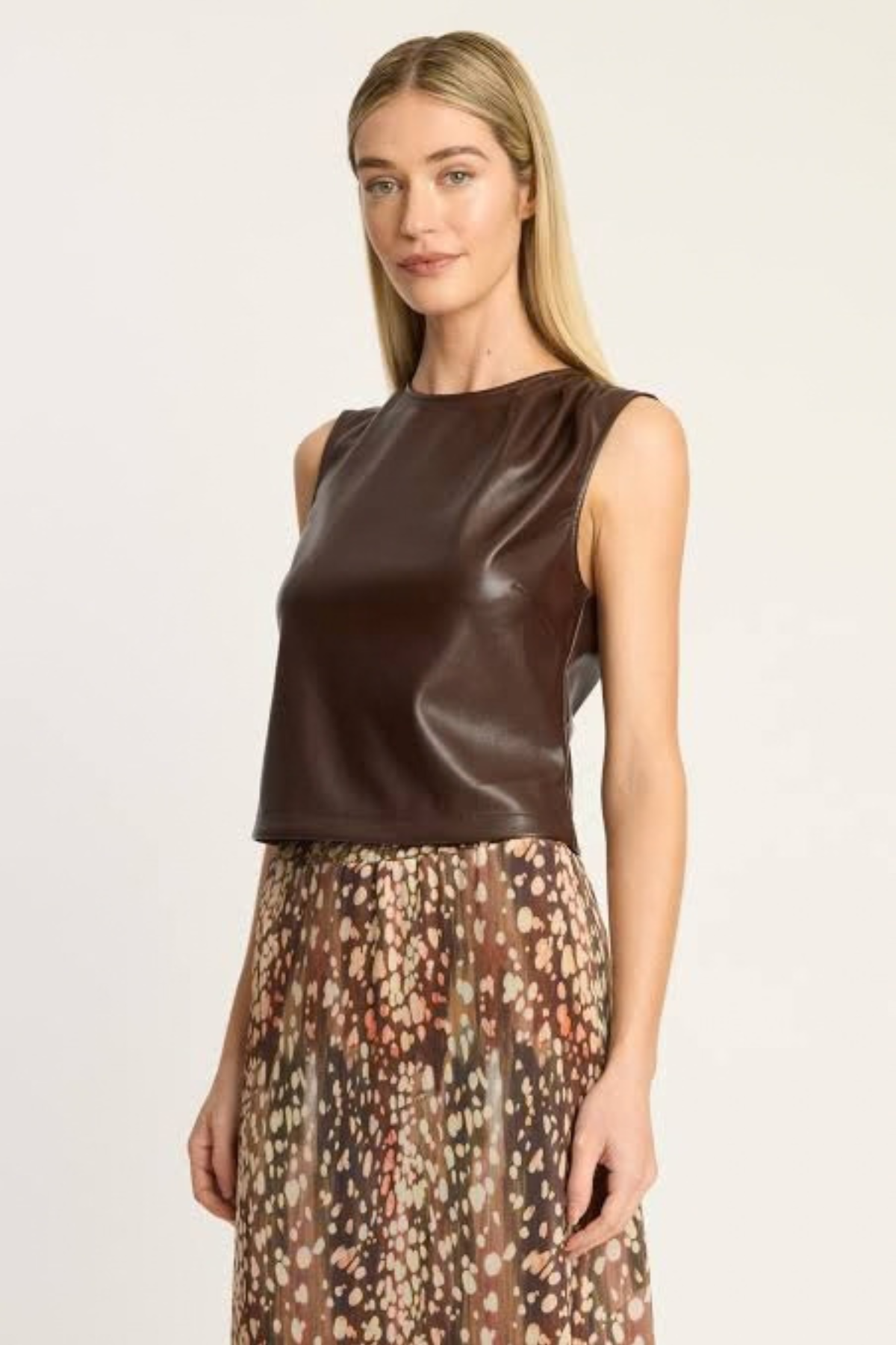 A woman modeling a sleeveless dark brown faux-leather top paired with a brown, beige, and pink patterned skirt, set against a plain light gray studio background.