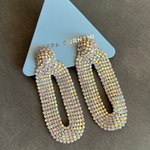 A pair of long, open-top dangle earrings covered in iridescent rhinestones, displayed on a light blue triangular card with the brand name 'GADA GURNANI' partially visible, set against a dark gray textured background.