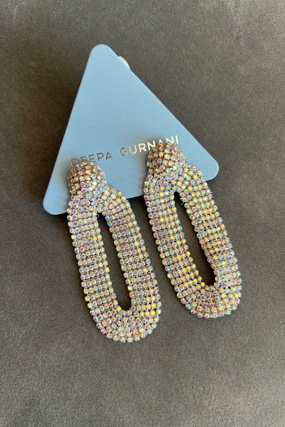 A pair of long, open-top dangle earrings covered in iridescent rhinestones, displayed on a light blue triangular card with the brand name 'GADA GURNANI' partially visible, set against a dark gray textured background.