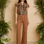 A woman stands in a studio styled with potted green plants, wearing a short-sleeve floral blouse and brown wide-leg cropped pants with brown heeled sandals.