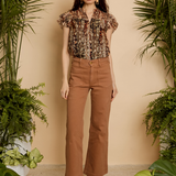 A woman stands in a studio styled with potted green plants, wearing a short-sleeve floral blouse and brown wide-leg cropped pants with brown heeled sandals.