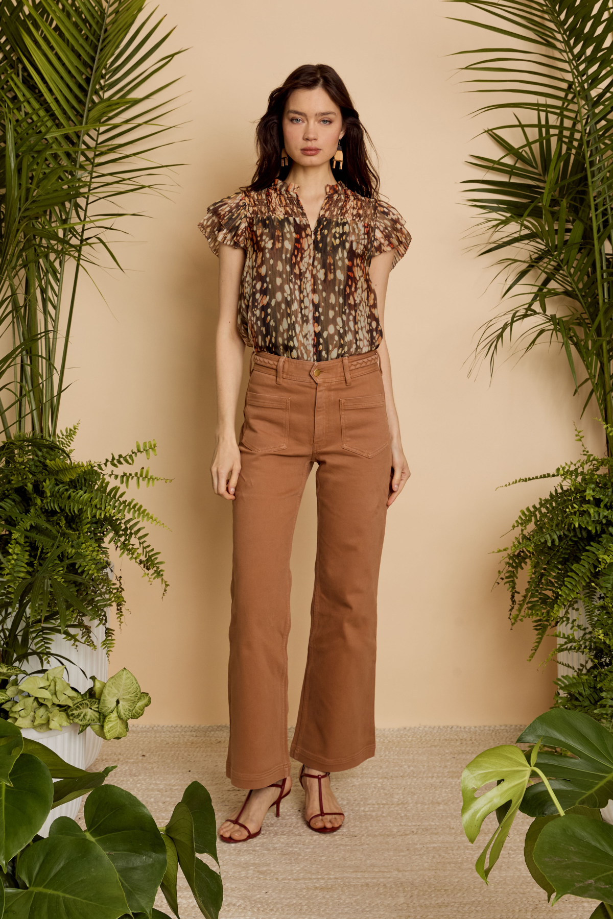 A woman stands in a studio styled with potted green plants, wearing a short-sleeve floral blouse and brown wide-leg cropped pants with brown heeled sandals.