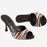 A pair of women's heeled slide sandals featuring a brown and white zebra print upper with dark brown straps and a dark brown stiletto heel, shown against a plain light gray background.