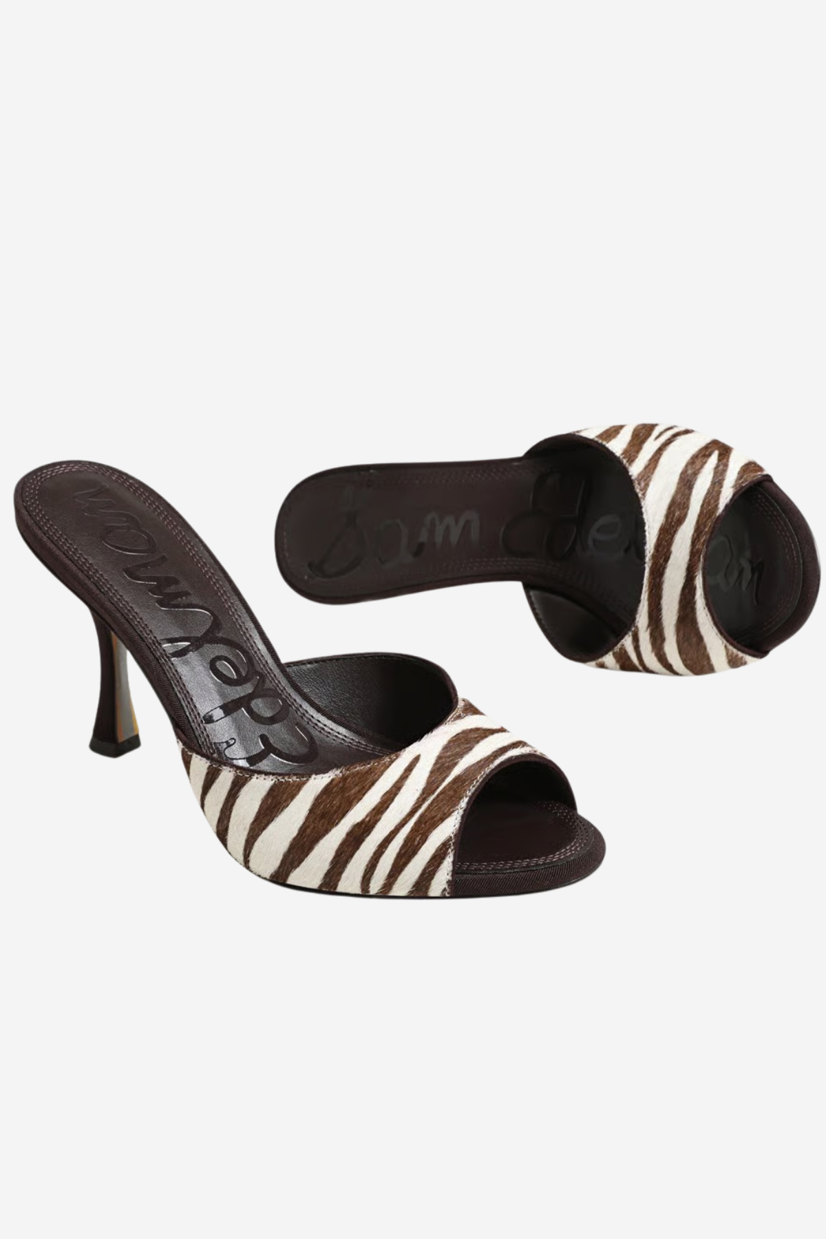 A pair of women's heeled slide sandals featuring a brown and white zebra print upper with dark brown straps and a dark brown stiletto heel, shown against a plain light gray background.