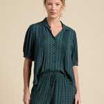 A woman modeling a dark green and black plaid short-sleeve button-up shirt and matching drawstring pants against a solid beige studio background.