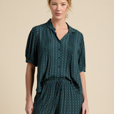 A woman modeling a dark green and black plaid short-sleeve button-up shirt and matching drawstring pants against a solid beige studio background.