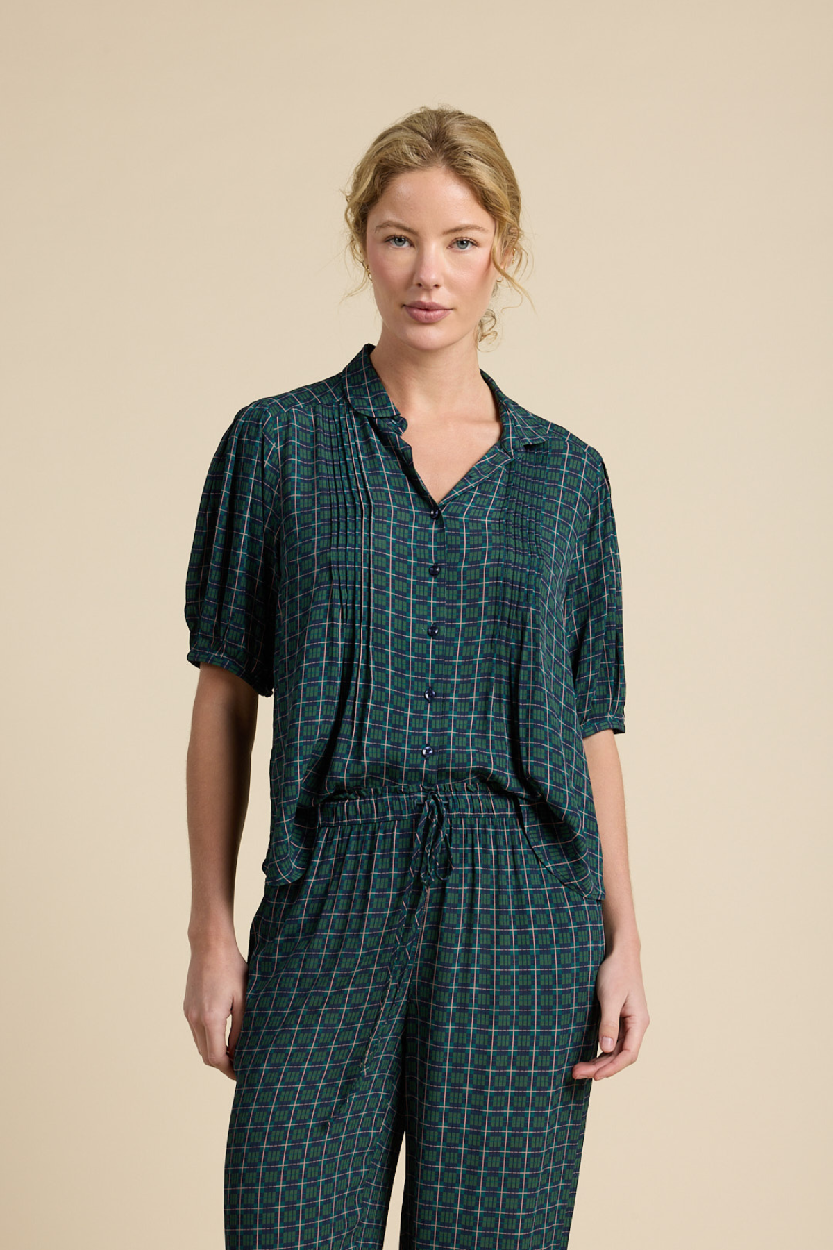 A woman modeling a dark green and black plaid short-sleeve button-up shirt and matching drawstring pants against a solid beige studio background.