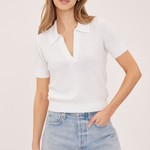 A woman modeling a white short-sleeve knit polo shirt with a split V-neckline, paired with light-wash denim shorts, against a solid light beige studio background.