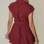 A woman shown from the back wearing a short burgundy dress with cap sleeves and a matching waist tie, against a plain beige studio background.