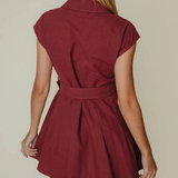 A woman shown from the back wearing a short burgundy dress with cap sleeves and a matching waist tie, against a plain beige studio background.
