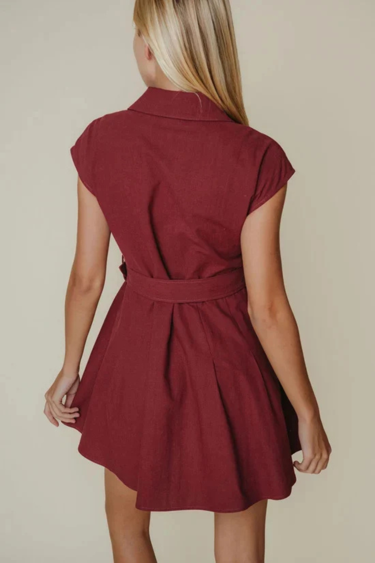 A woman shown from the back wearing a short burgundy dress with cap sleeves and a matching waist tie, against a plain beige studio background.