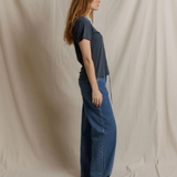 A woman with long light brown hair stands in profile wearing a dark slate blue short-sleeve t-shirt and wide-leg blue jeans, standing against a draped beige fabric backdrop.