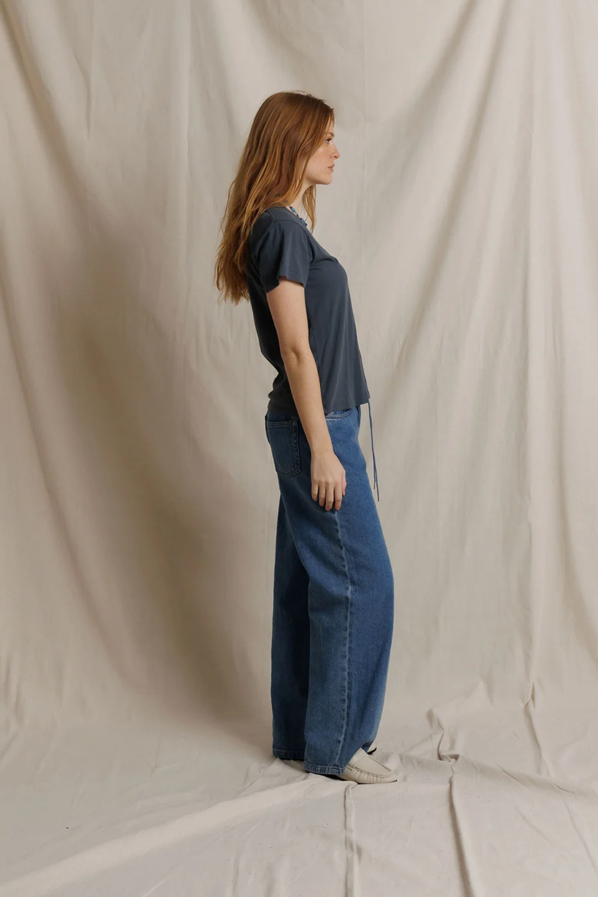 A woman with long light brown hair stands in profile wearing a dark slate blue short-sleeve t-shirt and wide-leg blue jeans, standing against a draped beige fabric backdrop.