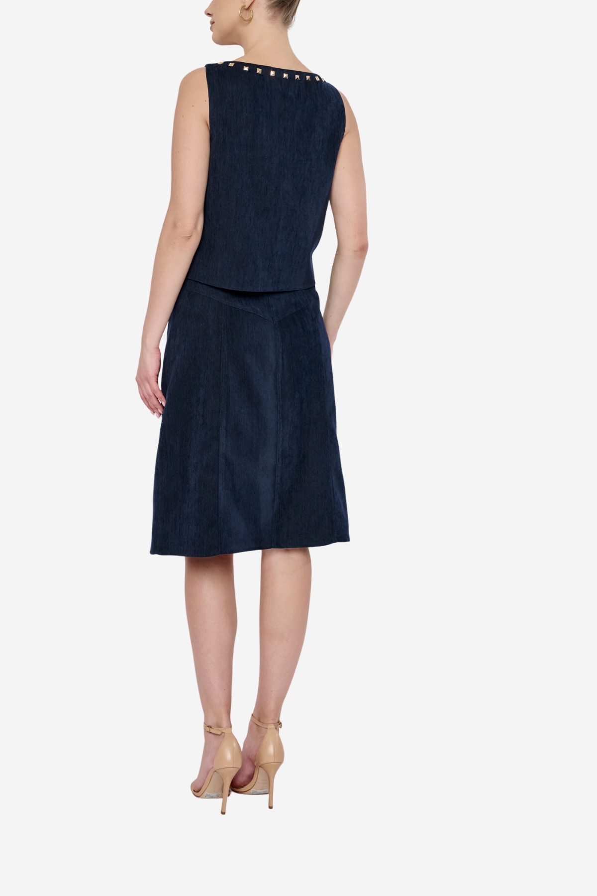 A woman shown from the back wearing a sleeveless navy blue dress with gold stud detailing around the neckline, paired with beige high-heeled sandals, against a light gray studio background.