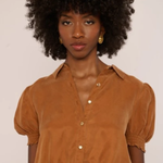 A woman with a large dark afro wearing a cropped, short-sleeved button-up blouse in a warm camel brown color, paired with matching brown bottoms. She is wearing small gold hoop earrings and posing against a plain off-white background.