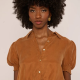 A woman with a large dark afro wearing a cropped, short-sleeved button-up blouse in a warm camel brown color, paired with matching brown bottoms. She is wearing small gold hoop earrings and posing against a plain off-white background.