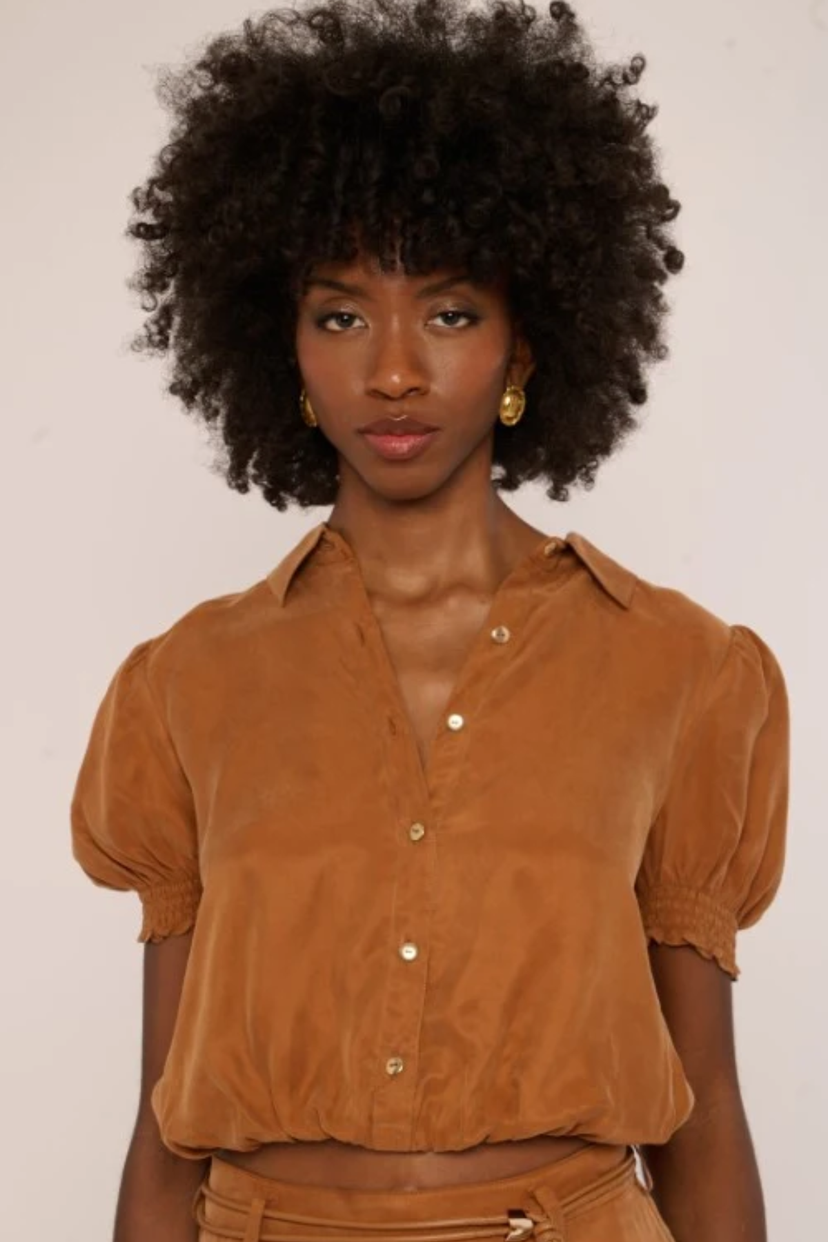A woman with a large dark afro wearing a cropped, short-sleeved button-up blouse in a warm camel brown color, paired with matching brown bottoms. She is wearing small gold hoop earrings and posing against a plain off-white background.