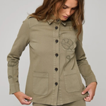 A woman wearing an olive green utility jacket with button-front closure, flap pockets, and a tonal embroidered chest motif, paired with matching olive pants, against a light gray studio background.