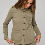 A woman wearing an olive green utility jacket with button-front closure, flap pockets, and a tonal embroidered chest motif, paired with matching olive pants, against a light gray studio background.
