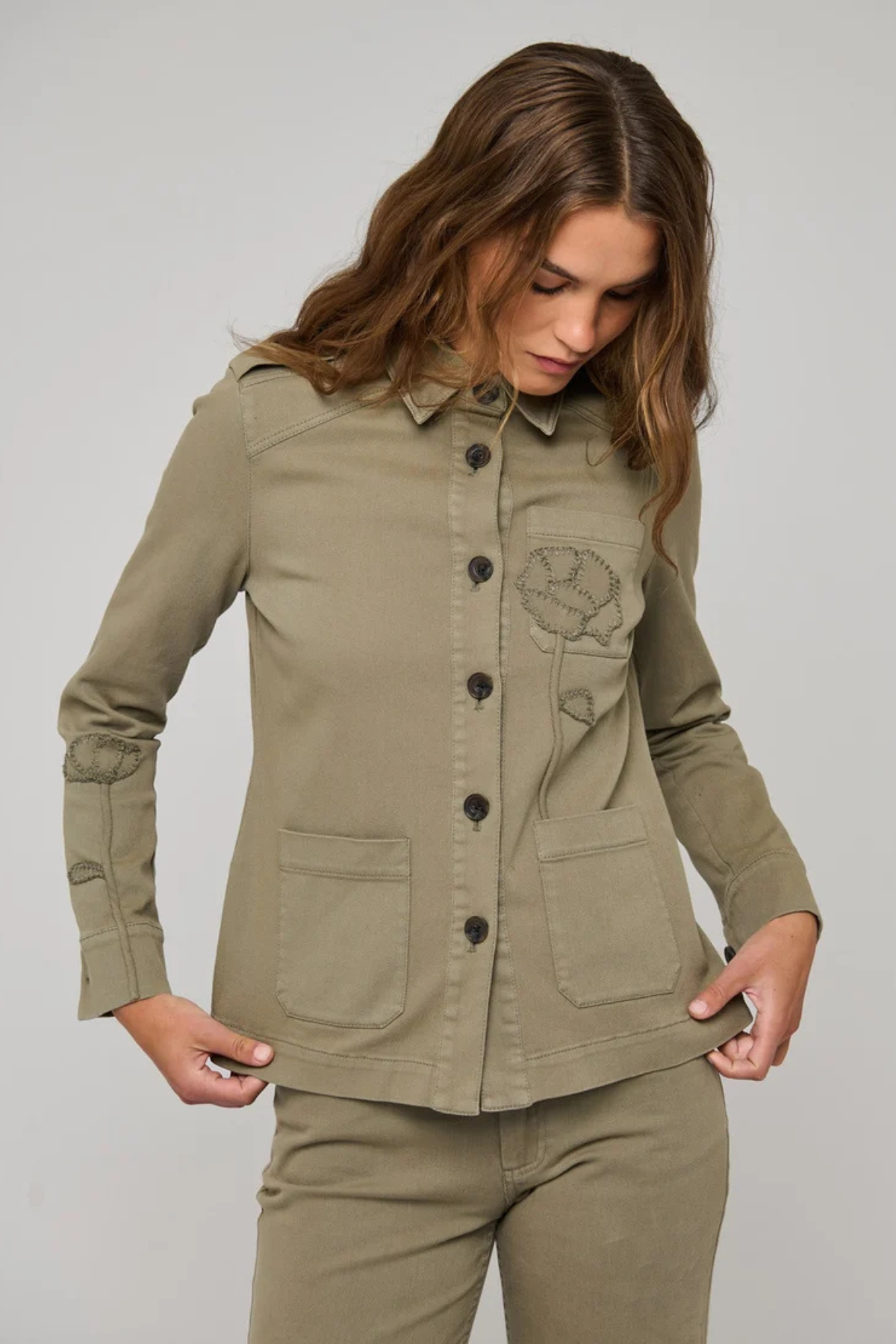 A woman wearing an olive green utility jacket with button-front closure, flap pockets, and a tonal embroidered chest motif, paired with matching olive pants, against a light gray studio background.