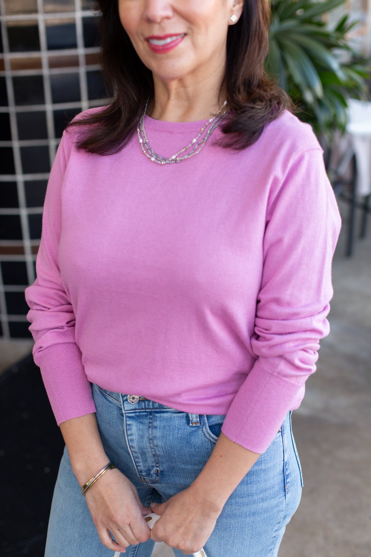 Woman wearing a pink sweater and blue jeans standing against a tiled wall.