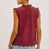 Woman wearing a burgundy sleeveless top with ruffle details and light blue jeans on a plain background